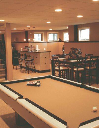 A well-lit basement recreation area with a pool table, dining set, and a kitchenette.