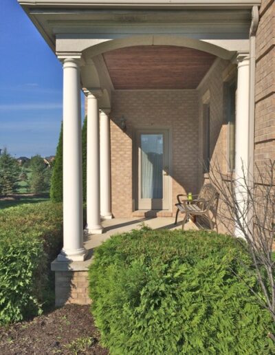 Front porch with columns and a bench, surrounded by green shrubbery.
