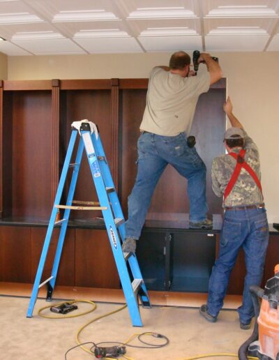 Two individuals installing cabinetry while using a ladder in an indoor setting.
