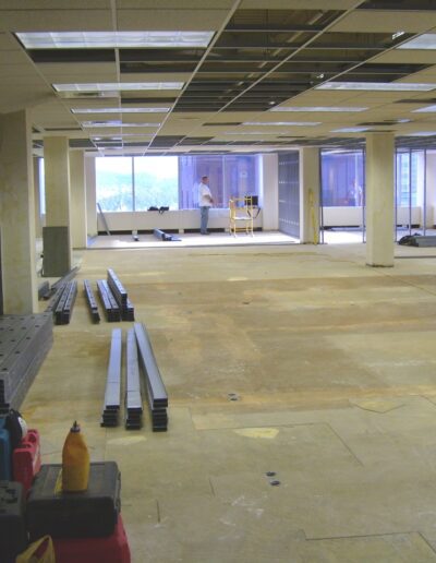 An unfinished office space under construction with exposed floors, materials stacked, and a worker in the background.