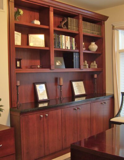 A well-organized wooden home office with a desk, bookshelf, and diplomas on the wall.
