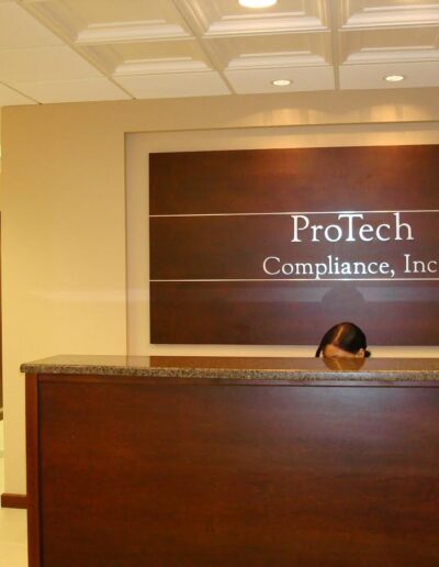 A reception area with the company sign "protech compliance, inc." above the desk, with only the receptionist's head visible.