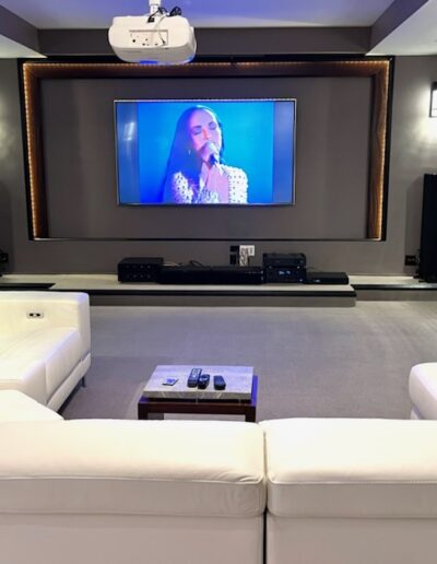 Modern home theater room with a large screen displaying a woman singing, flanked by speakers, and furnished with white sofas.