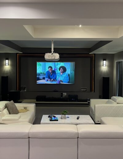 A home theater room with a large screen displaying a movie, surrounded by a white sectional sofa and dark walls.