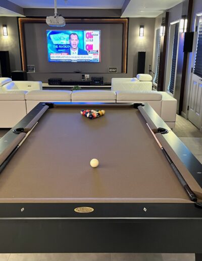 A modern home entertainment room with a pool table in the foreground and a television broadcasting news in the background.