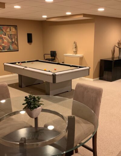 A modern basement recreation room with a pool table, seating area, and a workstation in the background.